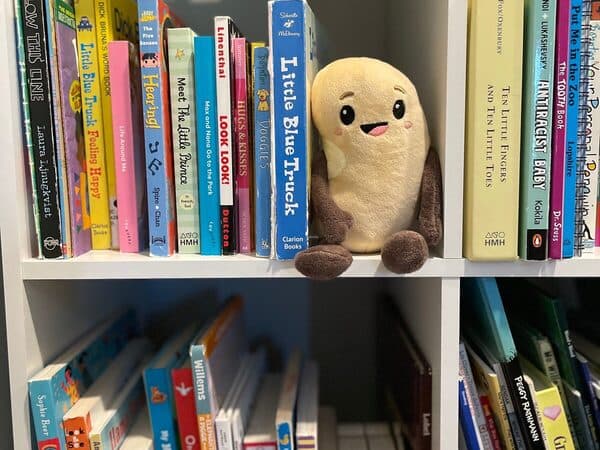 A Benny the Bean plushie sitting on a bookshelf of children’s books